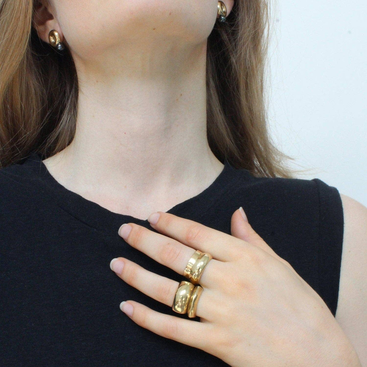 Woman wearing gold jewelry including earrings, rings, and a necklace on a plain background