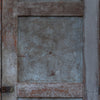 A close-up of a weathered wooden door panel with a central square section of distressed blue-gray metal or galvanized material, surrounded by peeling brown and gray paint.