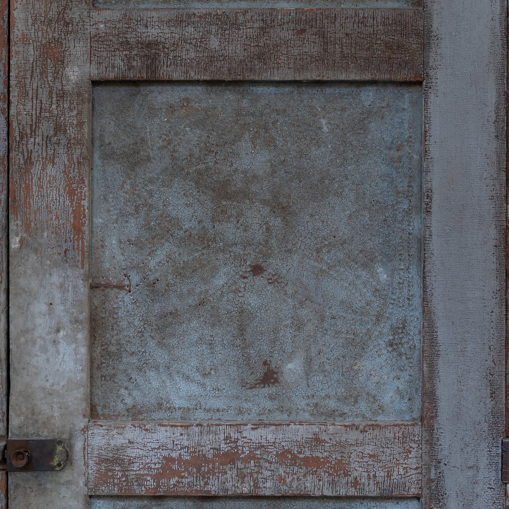A close-up of a weathered wooden door panel with a central square section of distressed blue-gray metal or galvanized material, surrounded by peeling brown and gray paint.