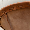 Close-up of a brown leather drum head with a stitched edge and eyelet holes, photographed against a light neutral background.