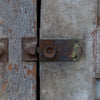 Close-up of a weathered metal latch and bolt on a distressed gray wooden door with peeling paint.