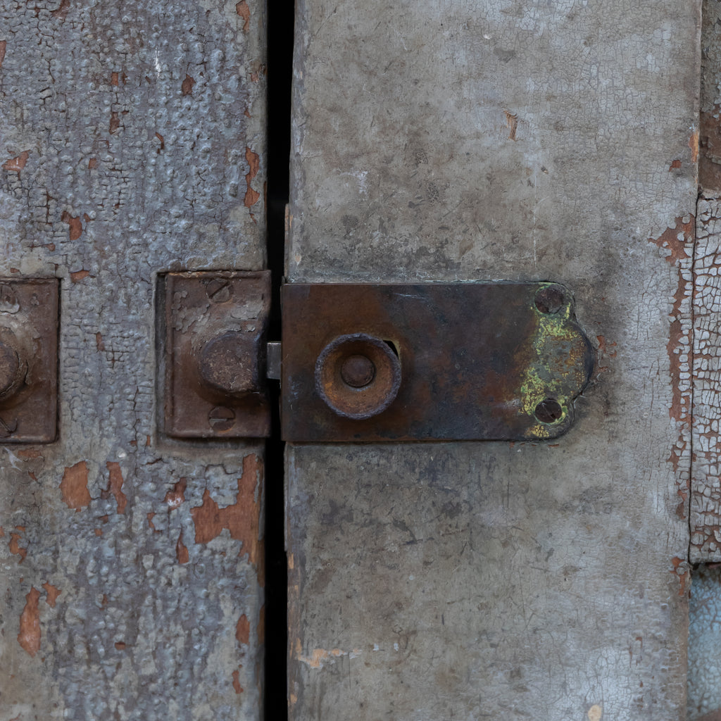 Close-up of a weathered metal latch and bolt on a distressed gray wooden door with peeling paint.