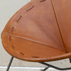 Close-up of a tan leather chair seat with decorative cutout stitching and a black metal frame, photographed against a light concrete wall.