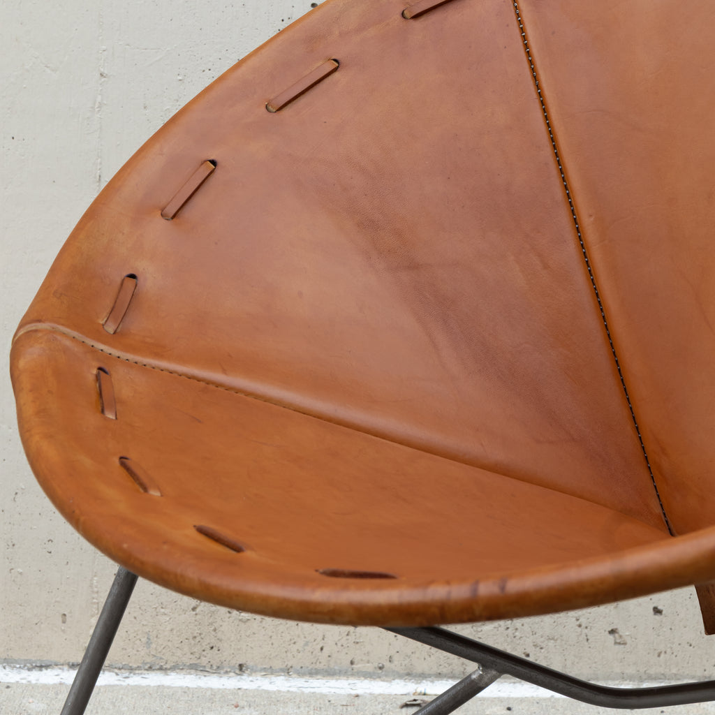 Close-up of a tan leather chair seat with decorative cutout stitching and a black metal frame, photographed against a light concrete wall.