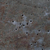 A close-up of a heavily textured, weathered surface with rusted brown and gray tones, featuring multiple small circular holes and a rough, metallic or concrete-like finish.
