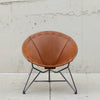 A mid-century modern accent chair with a warm brown leather seat and a black metal starburst-style leg base, set against a textured concrete wall.