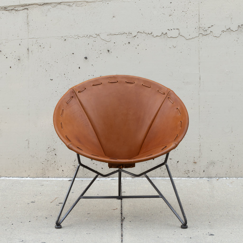 A mid-century modern accent chair with a warm brown leather seat and a black metal starburst-style leg base, set against a textured concrete wall.