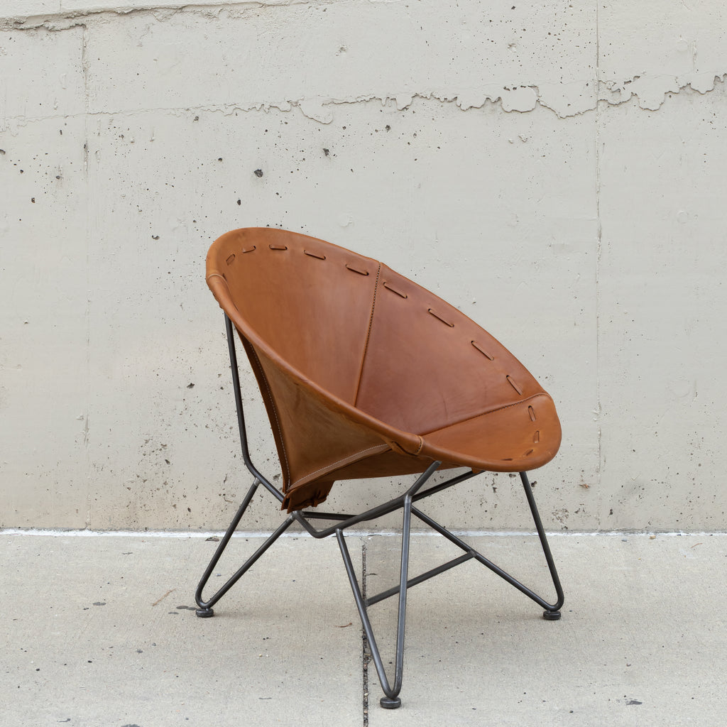 A mid-century modern spiral leather chair with a warm brown leather seat and a black metal tripod base, set against a textured concrete wall.