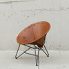 A mid-century modern spiral leather chair with a warm brown leather seat and black metal legs, set against a concrete wall and floor.