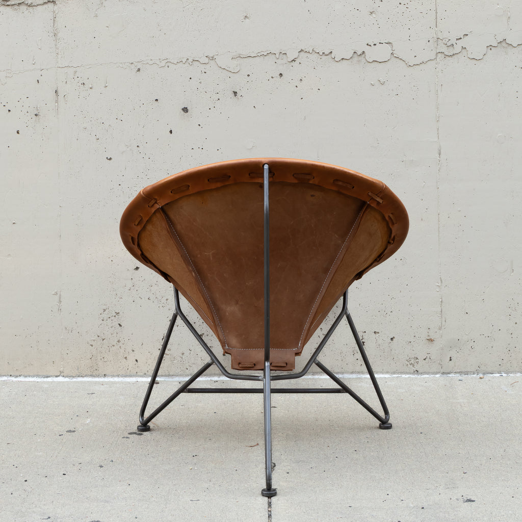 A mid-century modern spiral chair with a brown leather seat and backrest, supported by a black metal frame, photographed against a textured concrete wall.