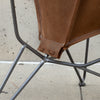 Close-up of a brown leather chair seat and back panel attached to a dark metal frame, photographed outdoors against a concrete wall and floor.