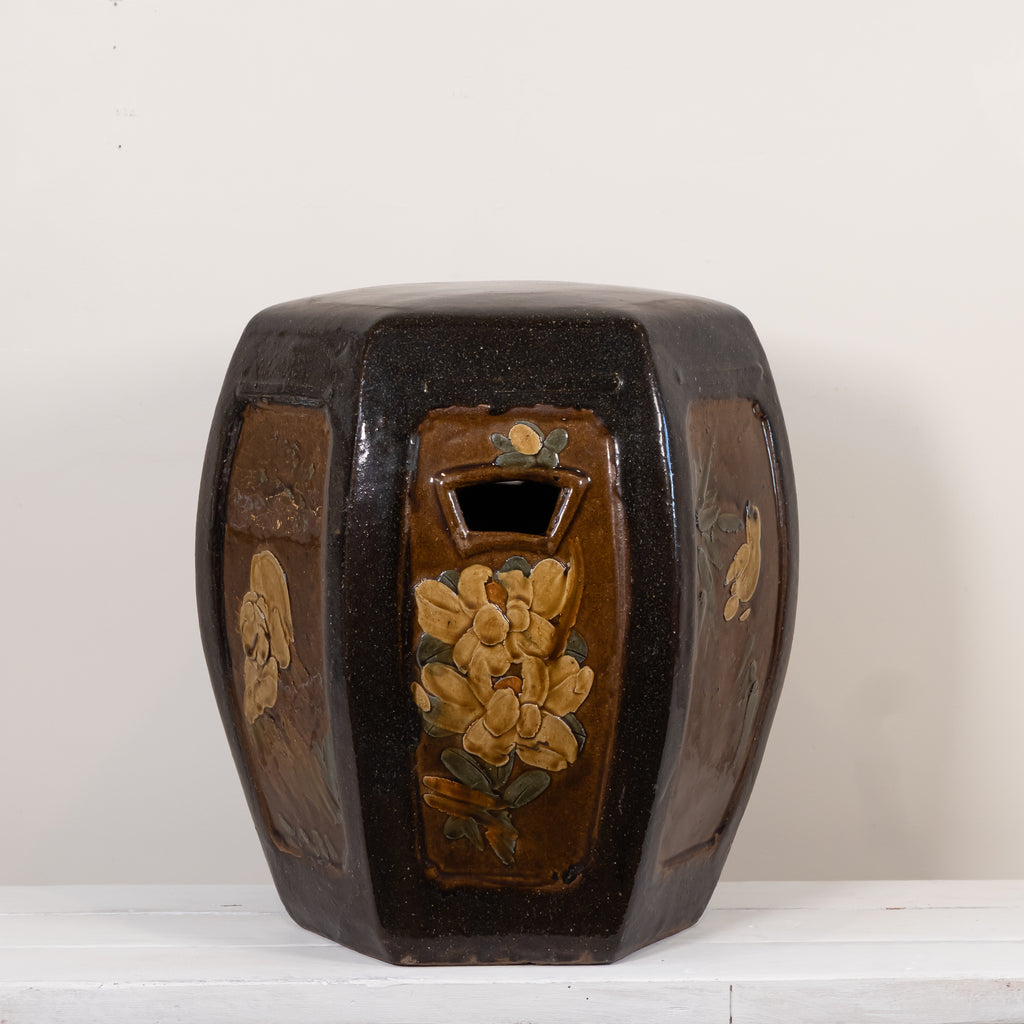 A vintage octagonal ceramic stool with a glossy dark brown glaze and hand-painted floral motifs in yellow, green, and orange, set against a plain off-white background.