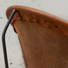 Close-up detail of a chair showing a curved brown leather seat, a matching leather backrest, and a black metal frame against a light concrete background.