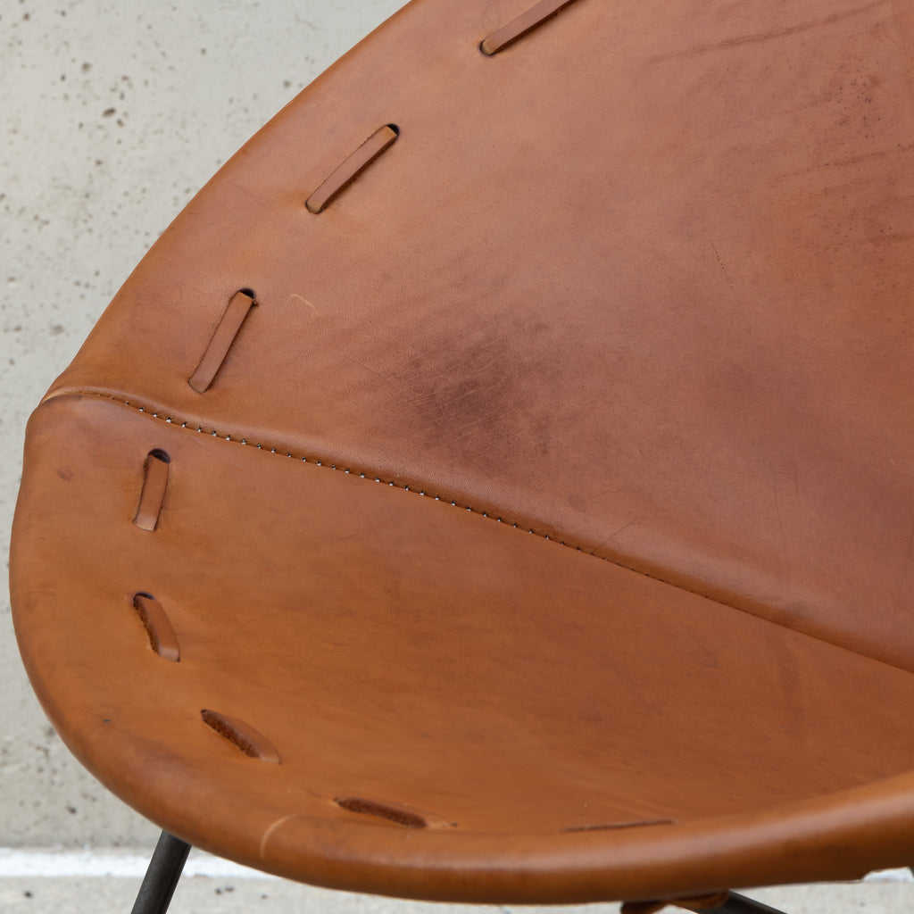 Close-up of a curved tan leather chair seat with stitched cutout details, photographed against a light concrete wall.