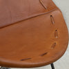 Close-up of a warm brown leather chair seat showing the texture, stitching, and seat hole construction.