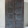 A rustic double-door cabinet or wardrobe made of weathered wood and distressed metal panels, featuring a weathered blue-gray finish with brown wood showing through, set against a plain off-white background.