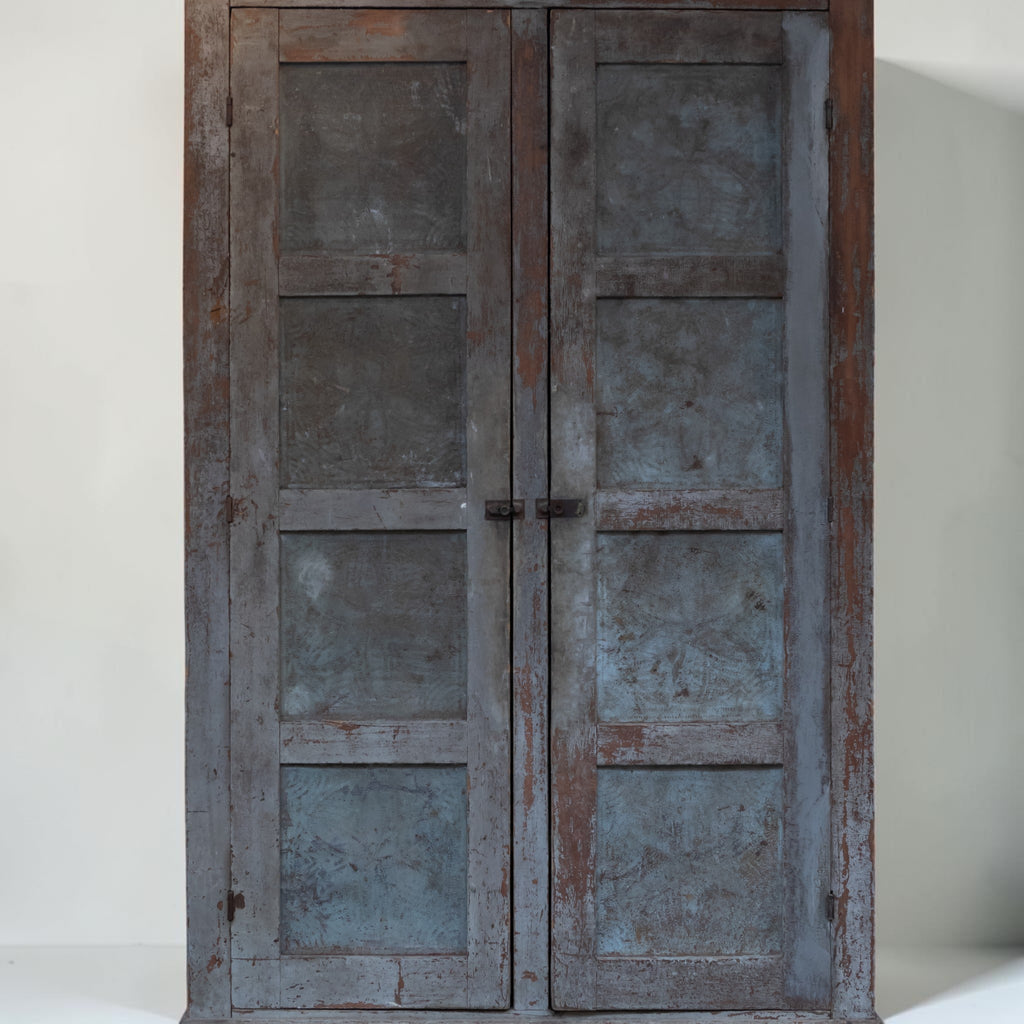 A rustic double-door cabinet or wardrobe made of weathered wood and distressed metal panels, featuring a weathered blue-gray finish with brown wood showing through, set against a plain off-white background.