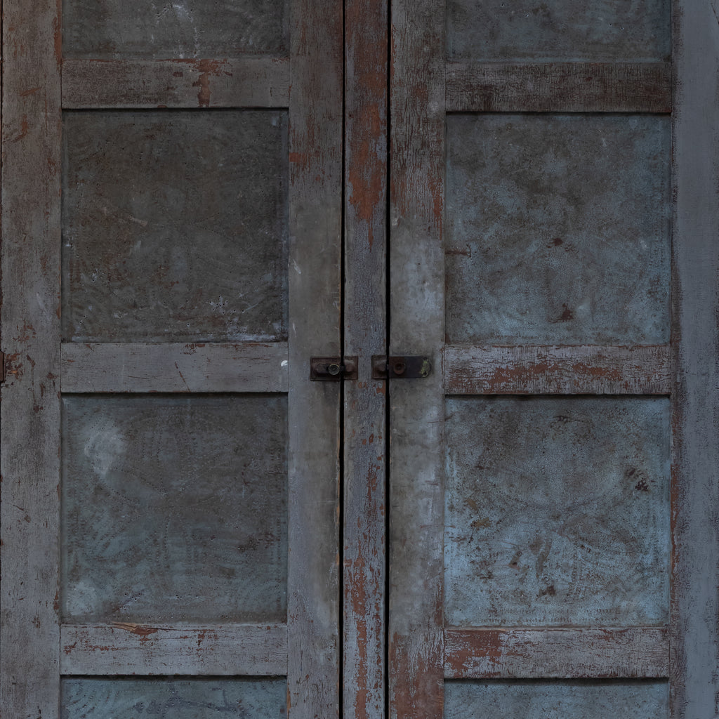 Rustic double door with weathered gray panels and peeling reddish-brown paint, secured by two dark metal latches in the center.