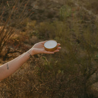 A hand holding a small round metal tin with a white lid, set against a blurred outdoor desert landscape with dry brush and warm sunlight.