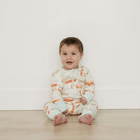 Baby sitting on a wooden floor wearing a patterned onesie against a white wall.