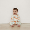 Baby sitting on a wooden floor wearing a patterned onesie against a white wall.
