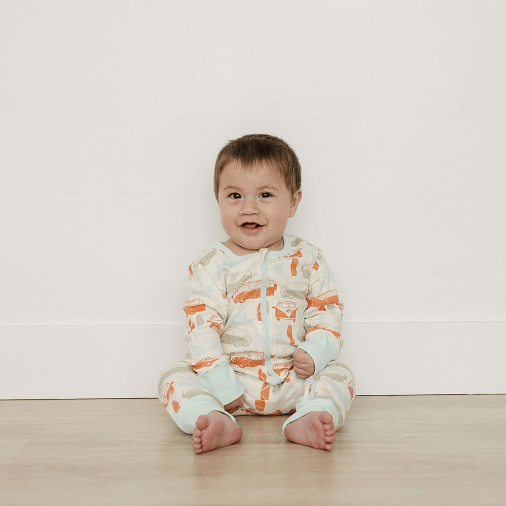 Baby sitting on a wooden floor wearing a patterned onesie against a white wall.