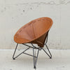 A mid-century modern leather chair with a warm tan leather seat and a black metal tripod base, set against a textured concrete wall.