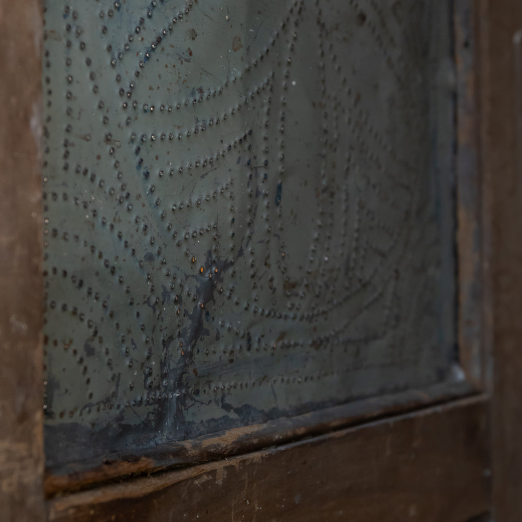 Close-up of a distressed, vintage-style metal door panel with a textured, embossed pattern of rivets on a weathered, dark gray surface, framed by a rustic brown wooden border.