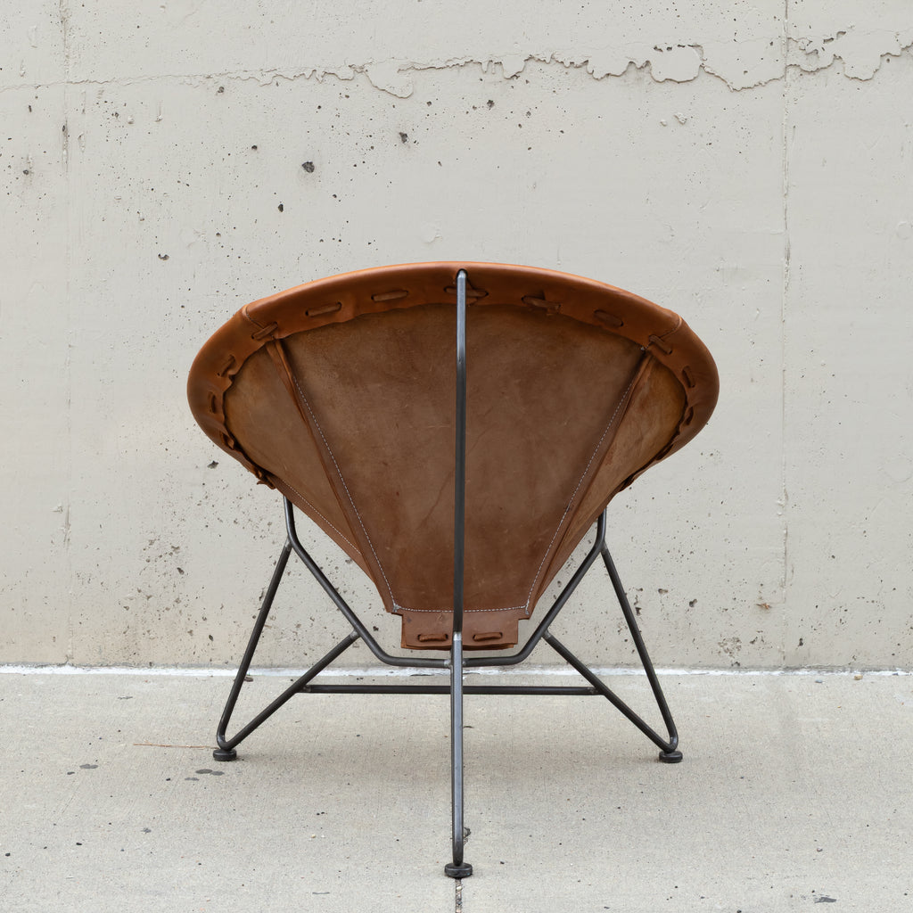 A mid-century modern spiral chair with a brown leather seat and backrest, supported by a black metal frame, photographed against a concrete wall.