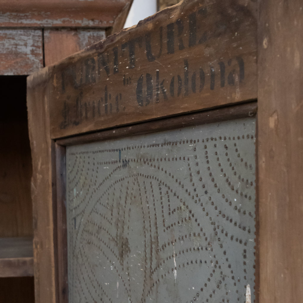 Close-up of a vintage wooden cabinet or screen with a metal patterned panel and stenciled text reading 'FURNITURE Liberty " Okolona"' on the top frame.