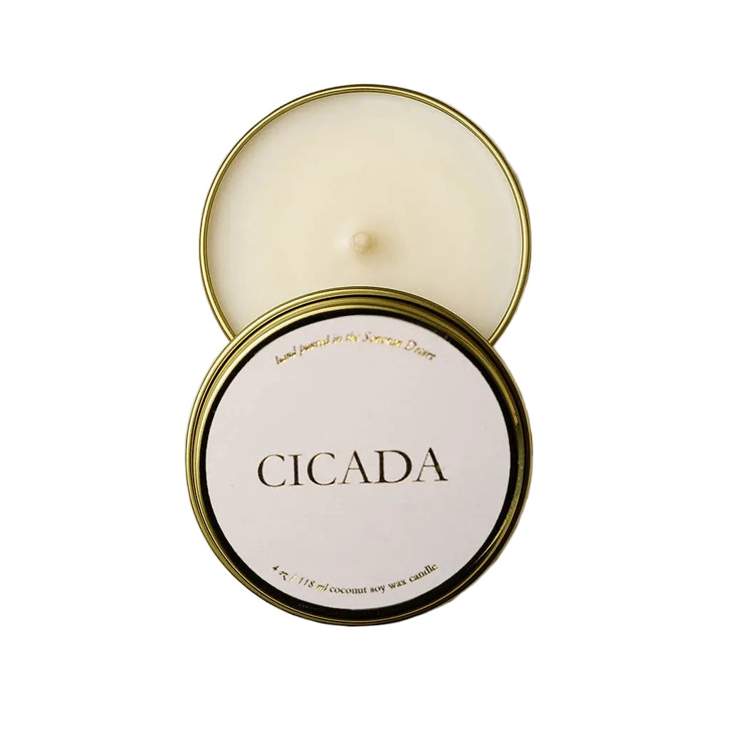 An open round metal candle tin showing cream-colored wax, with the lid placed on top. The front of the lid has a white label reading 'CICADA' in black serif text, with small gold text around the edge.