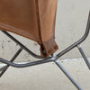 Close-up detail of a brown leather chair sling attached to a dark metal frame, showing the stitching and attachment point against a concrete wall.