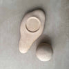 Two matte ceramic sculptural objects in a light warm beige tone, one organic teardrop-shaped piece with a circular recess and one smooth rounded sphere, arranged on a textured light gray surface.
