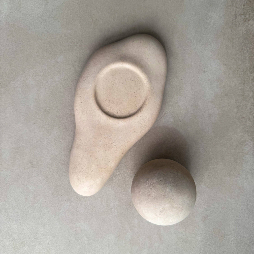 Two matte ceramic sculptural objects in a light warm beige tone, one organic teardrop-shaped piece with a circular recess and one smooth rounded sphere, arranged on a textured light gray surface.
