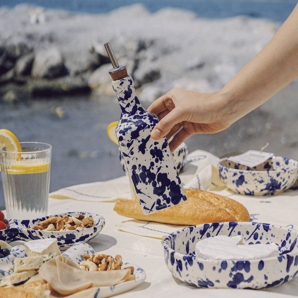 Person holding a floral-patterned bottle over a table with food and drinks by the ocean.