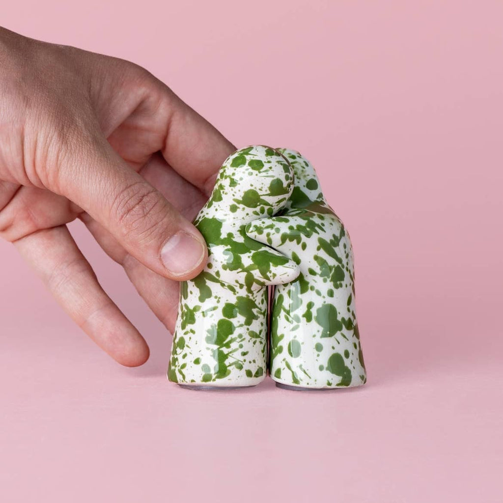 A hand holding a small ceramic figurine shaped like a kneeling figure with a green and white speckled pattern, set against a solid pink background.