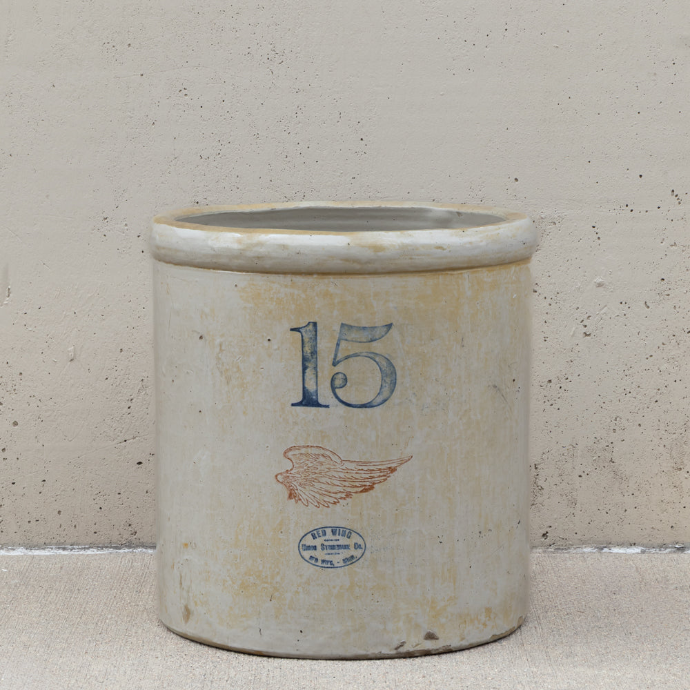 An antique 15 oz stoneware crockery jar with a distressed off-white glaze, featuring a blue printed number 15, a brown wing graphic, and a blue circular stamp reading 'New Orleans Co. 1870'.