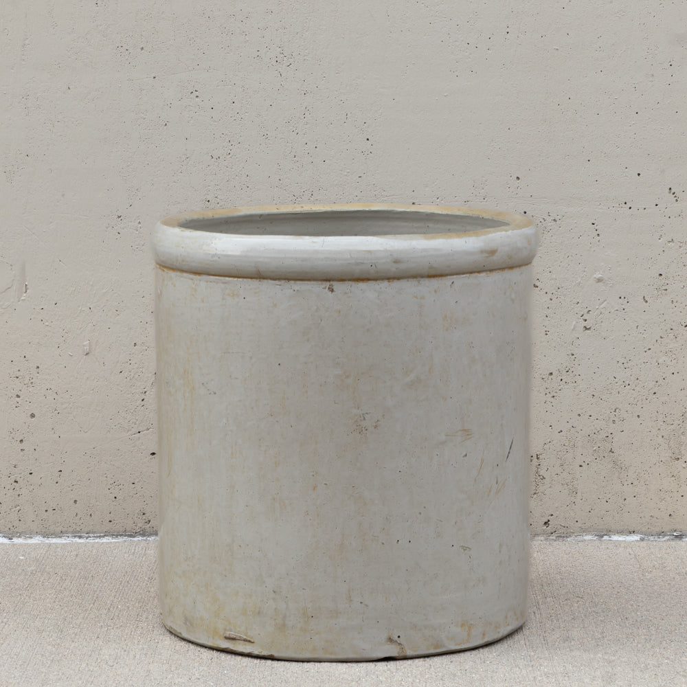 A cylindrical ceramic pot or planter with a wide rim, shown on a textured beige floor against a matching beige wall.