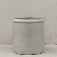 A cylindrical ceramic pot or planter with a wide rim, shown on a textured beige floor against a matching beige wall.