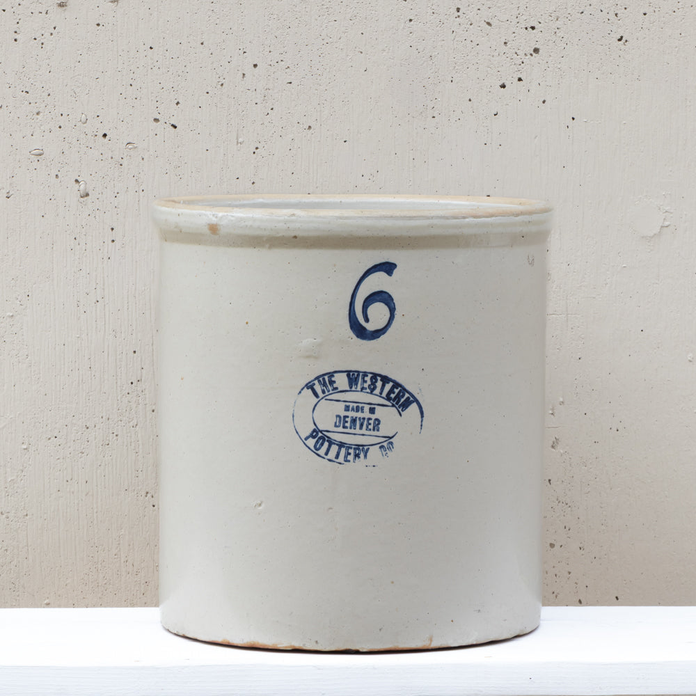 A vintage white stoneware crock pot with blue printed markings, including a blue number 6 and a circular logo for 'The Western Denver Pottery', set against a textured off-white background.