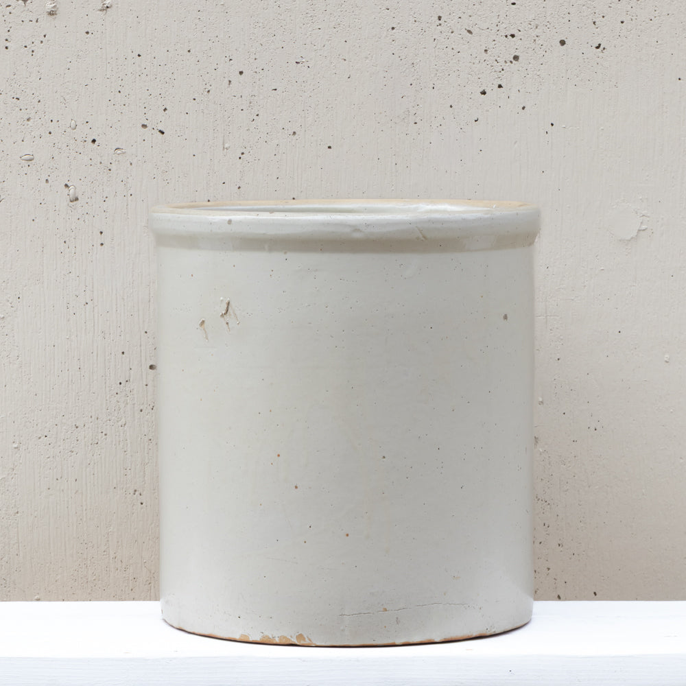 A tall, cylindrical ceramic stoneware container with a matte off-white glaze and a slightly flared rim, set against a textured beige plaster wall.