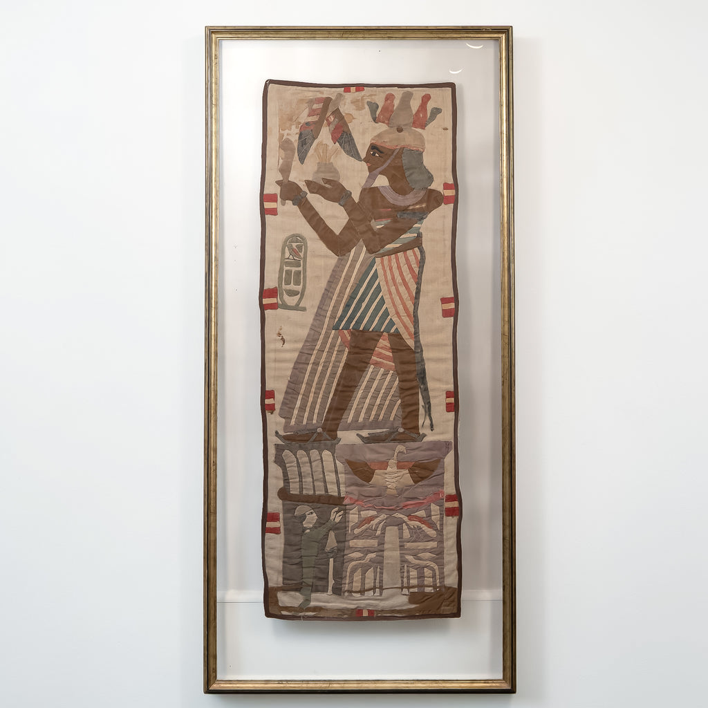 An Egyptian-style framed textile artwork featuring a stylized figure in profile, mounted in a clear glass frame with a gold-toned border against a plain white wall.