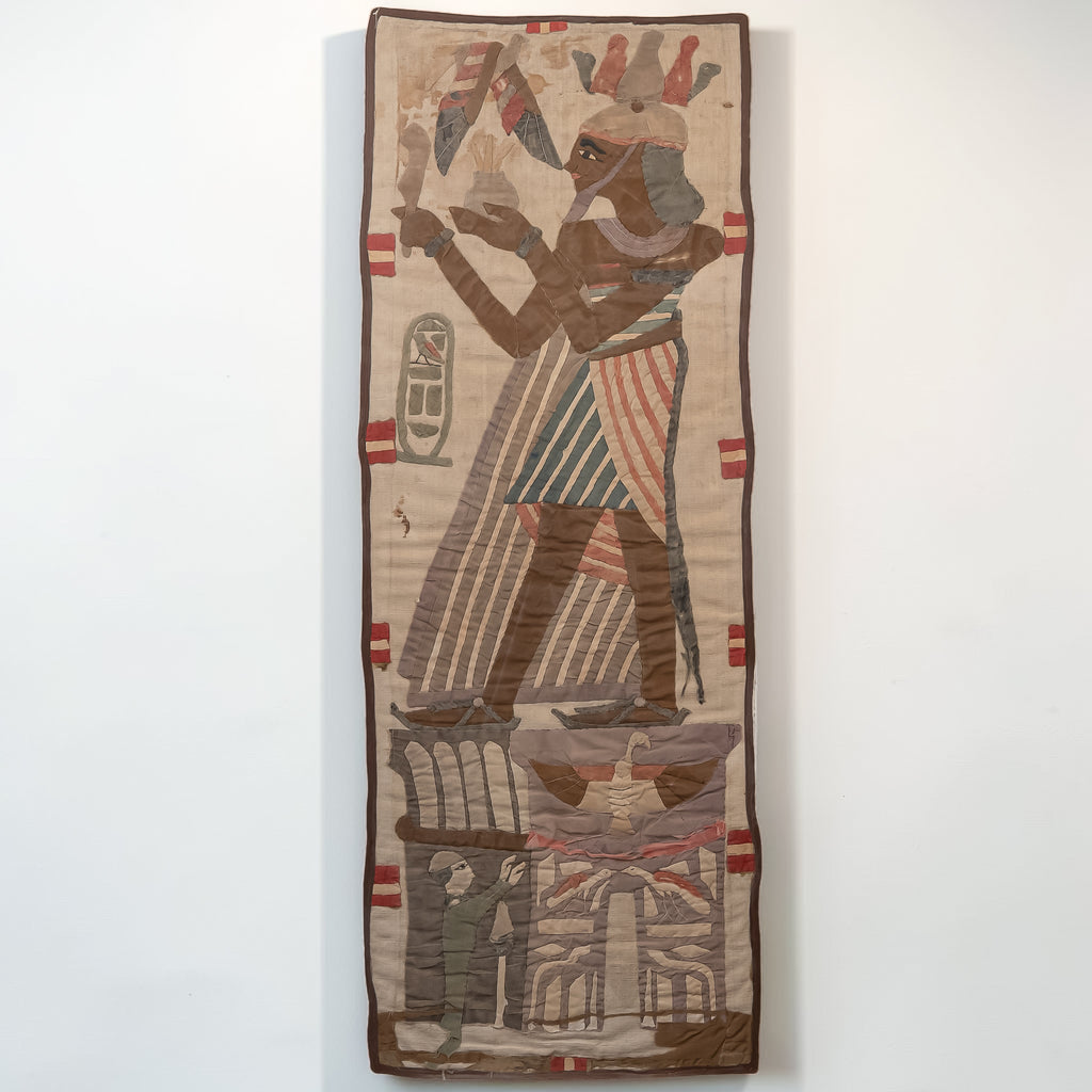 An Egyptian-style woven wall hanging or tapestry featuring a standing figure in profile with striped garments and headdress, set against a beige background with brown, red, and blue details, displayed against a plain white wall.