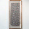 A framed textile art piece featuring a vertical rectangular panel with an intricate floral pattern in blue, pink, and beige, enclosed in a clear glass frame with a light wood border, mounted on a white wall.