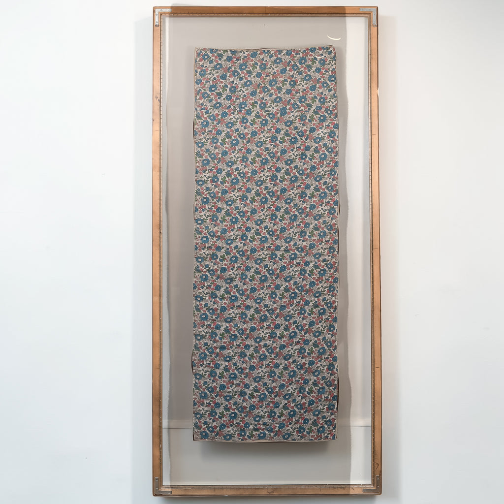 A framed textile art piece featuring a vertical rectangular panel with an intricate floral pattern in blue, pink, and beige, enclosed in a clear glass frame with a light wood border, mounted on a white wall.