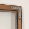 Close-up of a rustic brown wooden picture frame corner with a silver metal L-bracket and hanging ring against a plain off-white wall.