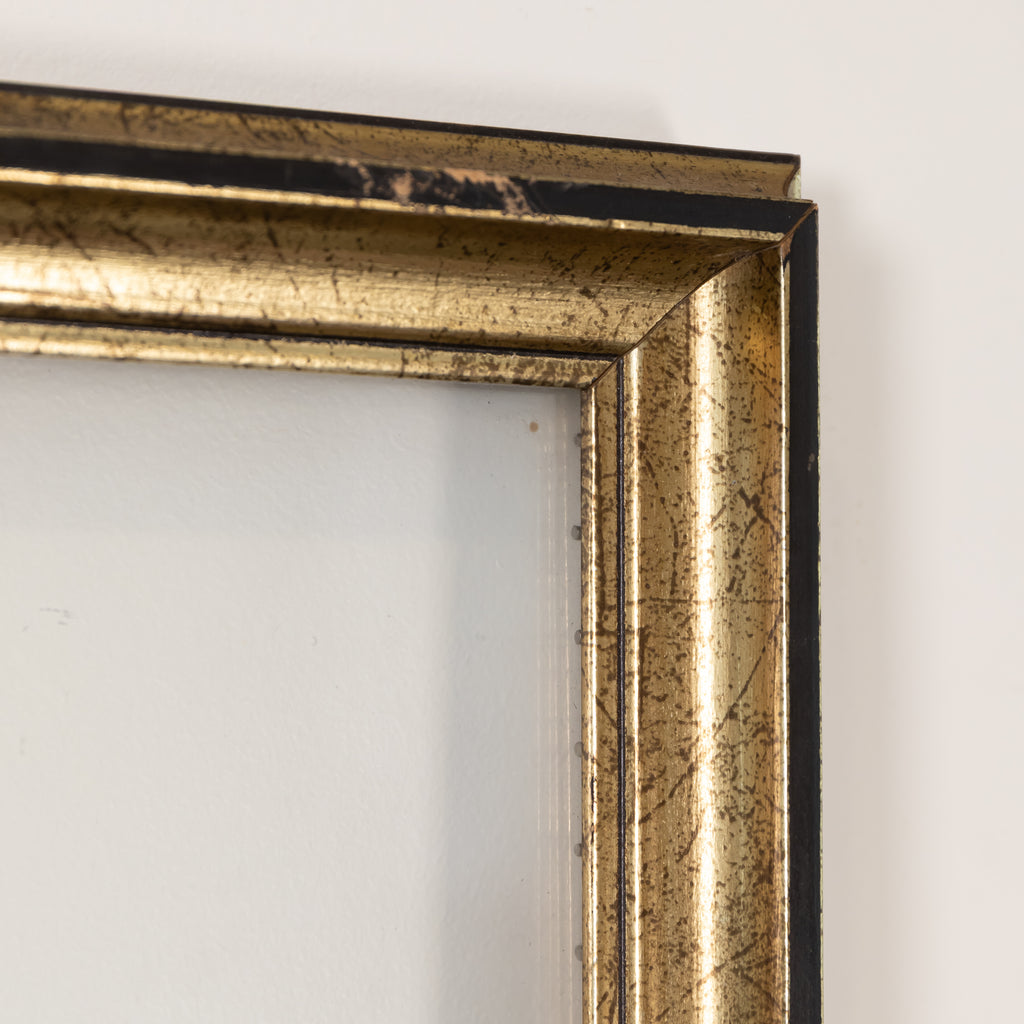 Close-up of a distressed gold picture frame corner with black distressing, set against a plain white wall.
