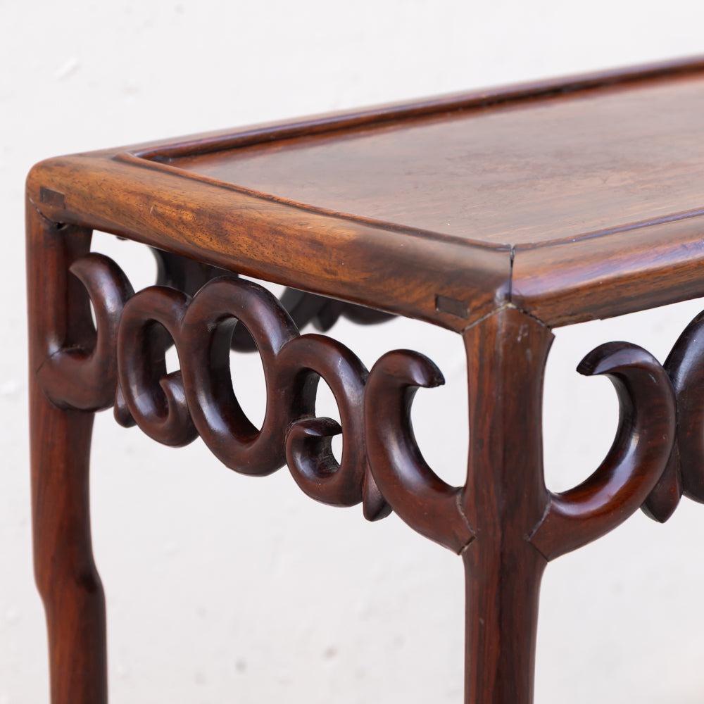 Close-up of a dark brown wooden table or console table leg and apron with carved scrollwork details, photographed against a plain light background.