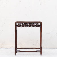 A dark brown wooden side table or stand with an ornate carved scrollwork apron and open lower support, photographed against a light textured wall.
