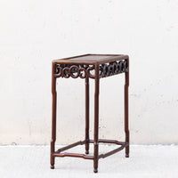 A dark brown wooden side table with an ornate openwork scrollwork apron and four straight legs, set against a textured off-white wall.